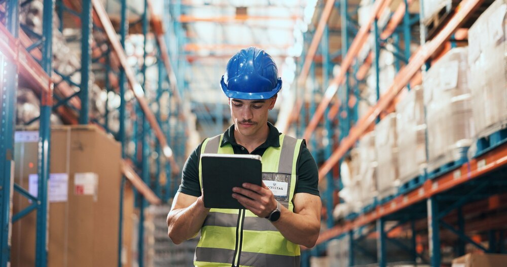 Operador de colete e capacete azul consultando um tablet entre as estantes de um armazém