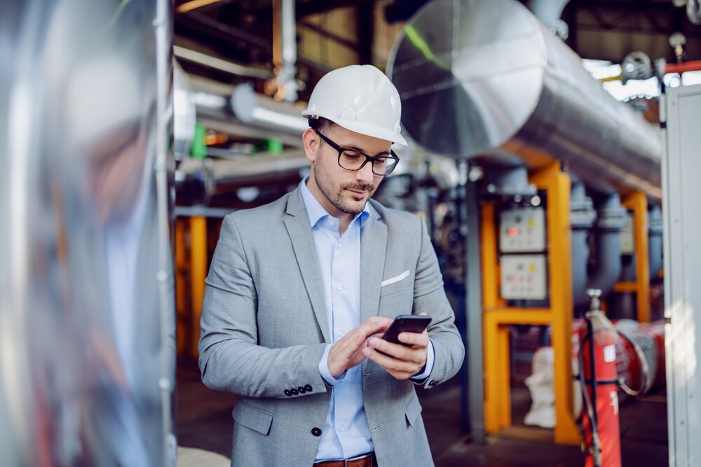 Homem de terno e capacete branco usando o celular numa planta industrial